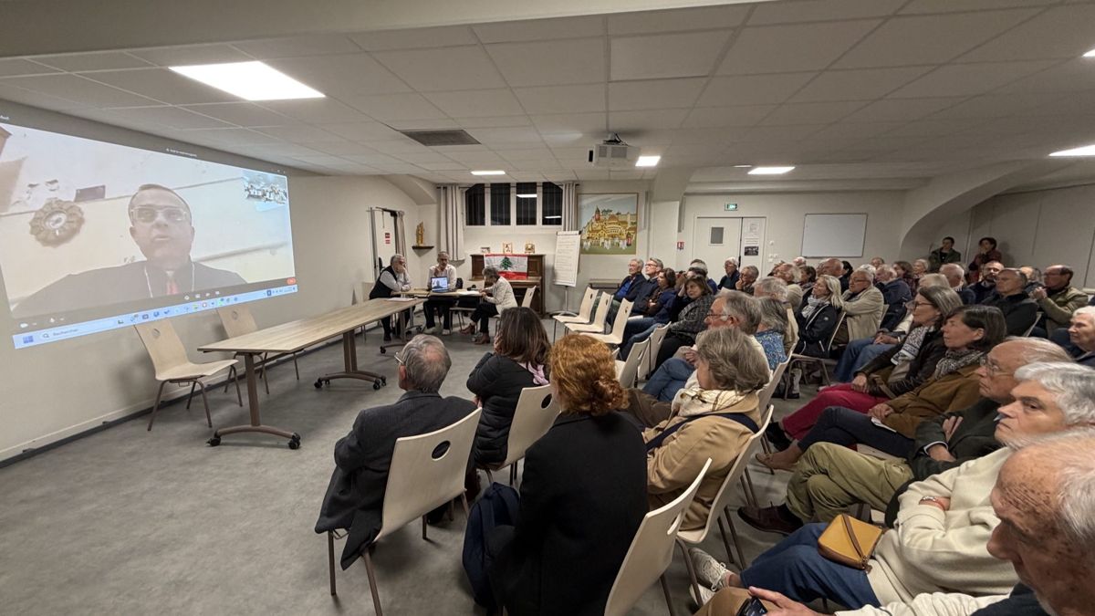Une salle paroissiale remplie de paroissiens face à un grand écran où apparaît Mgr Charbel Abdallah, archevêque maronite de Tyr.