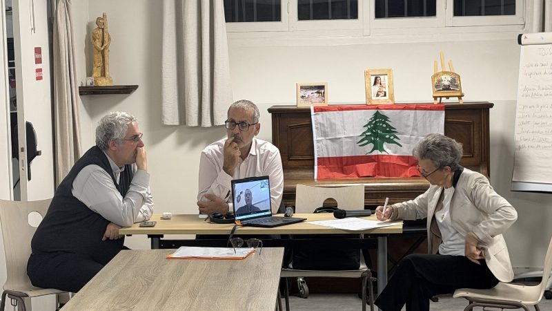 Trois personnes assises à une table devant un ordinateur portable où apparaît l'archevêque ; un drapeau libanais est posé sur un piano derrière elles.